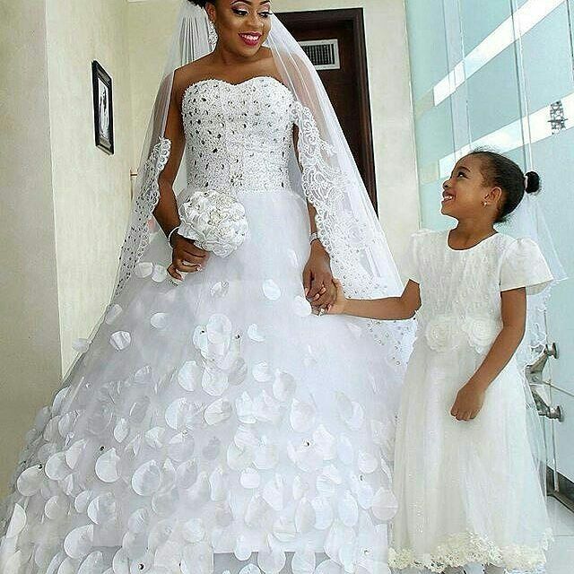 Woman in a white strapless wedding gown with floral appliqués and a long veil, holding a bouquet, alongside a girl in a white dress with a floral detail