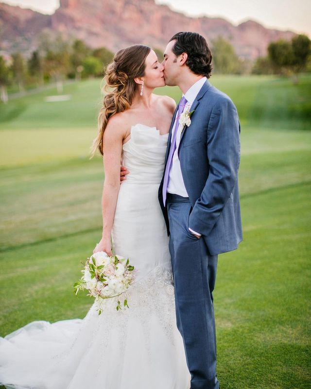 Woman in a white strapless wedding gown with lace detailing, holding a bouquet of white flowers, standing next to a man in a grey suit