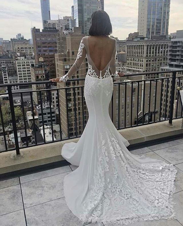 Woman in a white lace mermaid gown with long sleeves and an open back, standing on a balcony overlooking the city