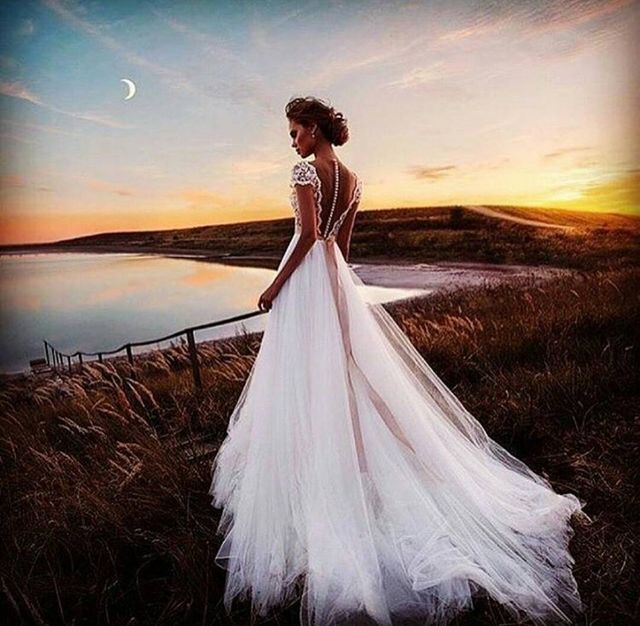 Woman in a white tulle wedding gown with a low back and lace cap sleeves, standing by a lake at sunset