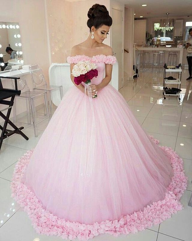 Woman in a pink off-the-shoulder ball gown with a tulle skirt and floral embellishments, holding a bouquet of flowers