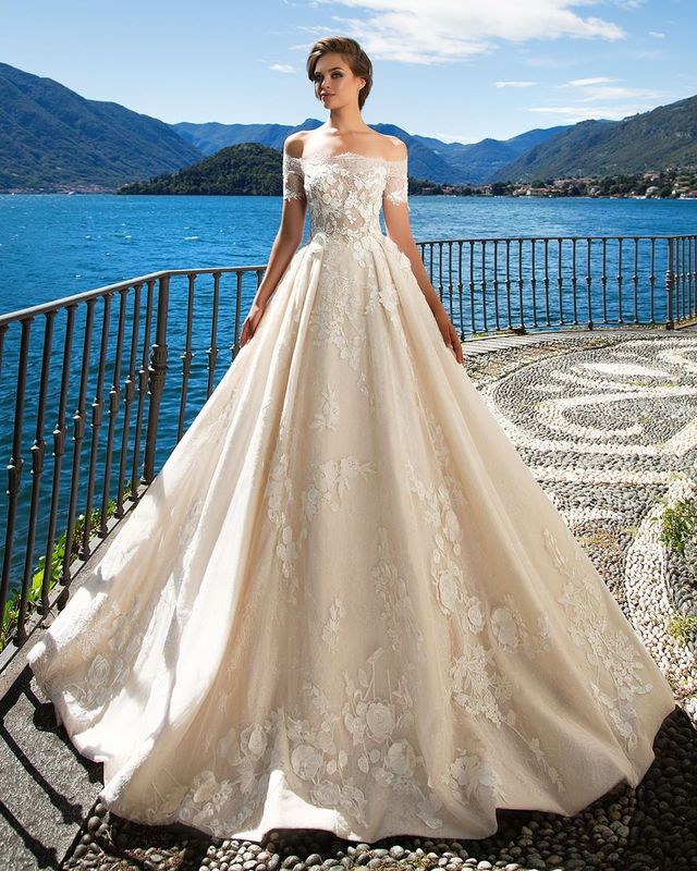 Woman in an off-the-shoulder ivory ball gown with floral embroidery and a full skirt by a lake