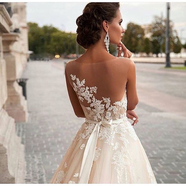 Woman in a cream lace wedding dress with a sheer back and a satin ribbon belt, wearing drop earrings