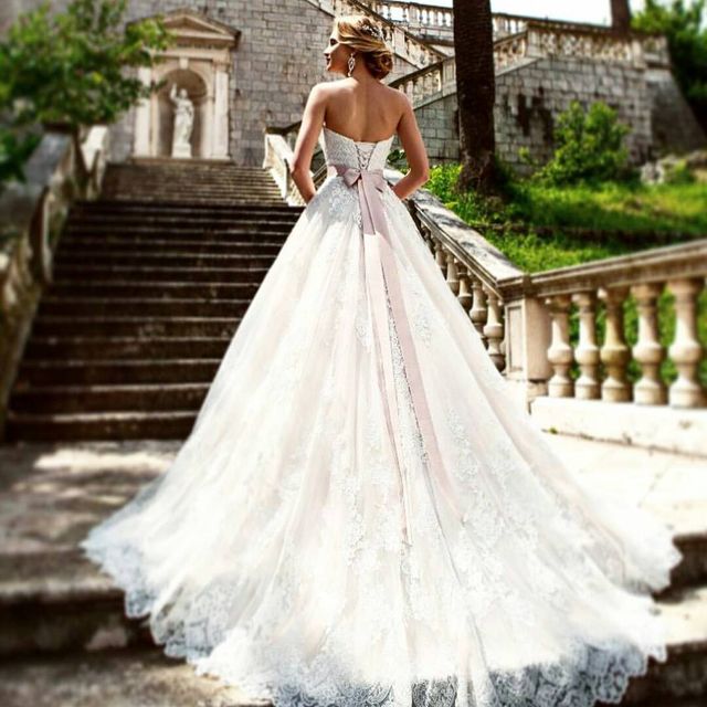 Woman in a white lace wedding gown with a pink satin bow, standing on stone steps