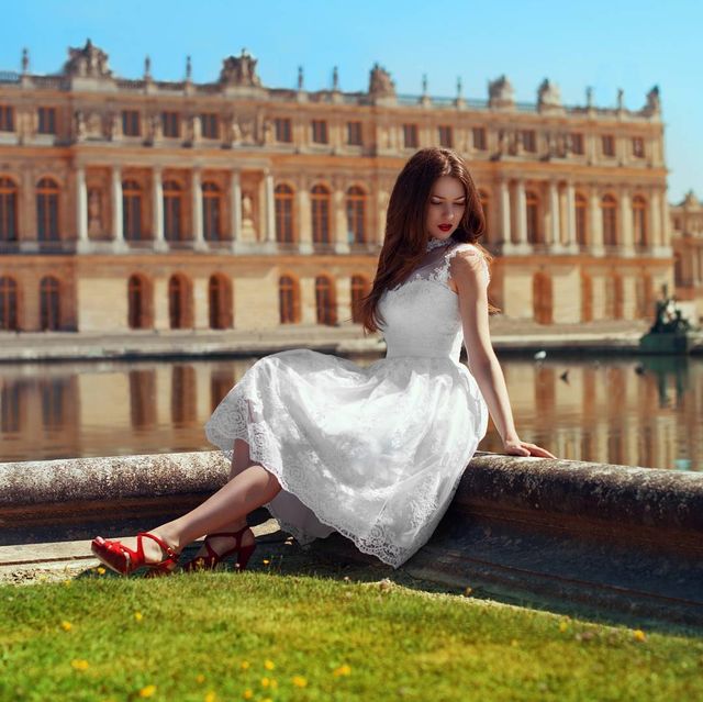 Woman in a white lace midi dress and red heeled sandals by a body of water