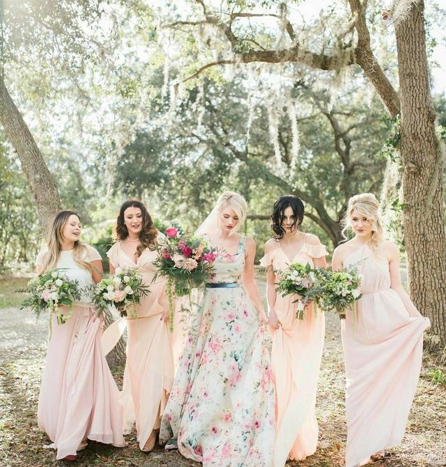 Woman in a floral ball gown with a blue sash, surrounded by women in pale pink maxi dresses, all holding bouquets