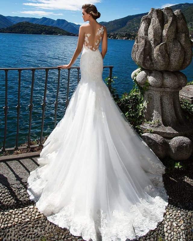 Woman in a white lace mermaid wedding gown with a sheer back and flowing train by a lake