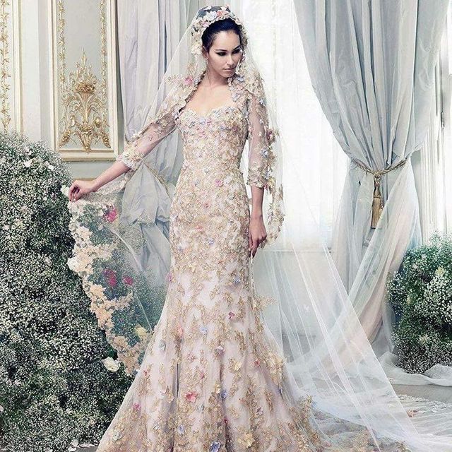Woman in a light pink embroidered wedding gown with a fitted silhouette and long lace sleeves, wearing a matching veil