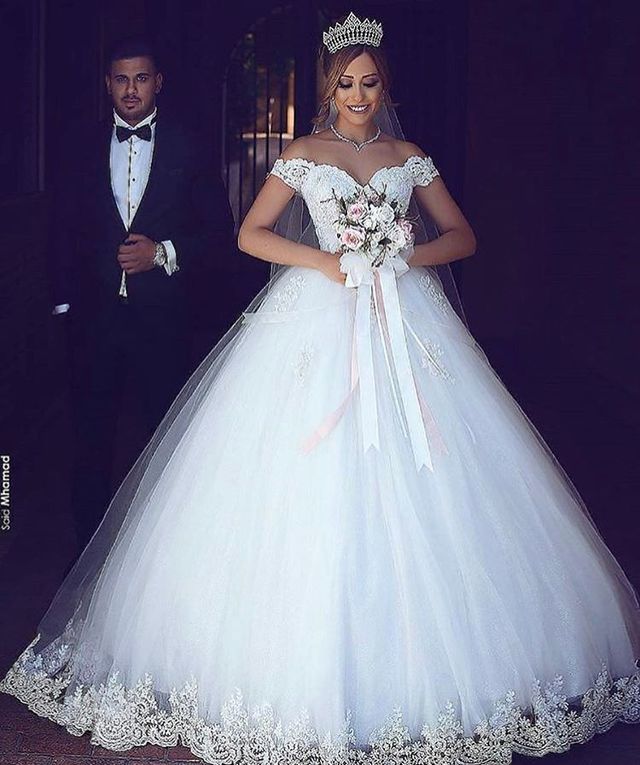 Woman in a white off-the-shoulder ball gown with lace detailing and a long veil, holding a bouquet of flowers
