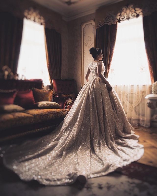 Woman in a sparkling silver ball gown with a long train and fitted bodice, standing by a window