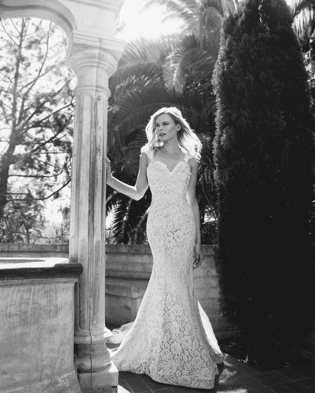 Woman in a white lace wedding gown with a fitted silhouette and cap sleeves, standing by a stone column