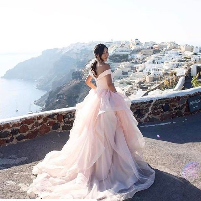 Woman in a pink tulle ball gown with off-the-shoulder sleeves, standing against a scenic backdrop