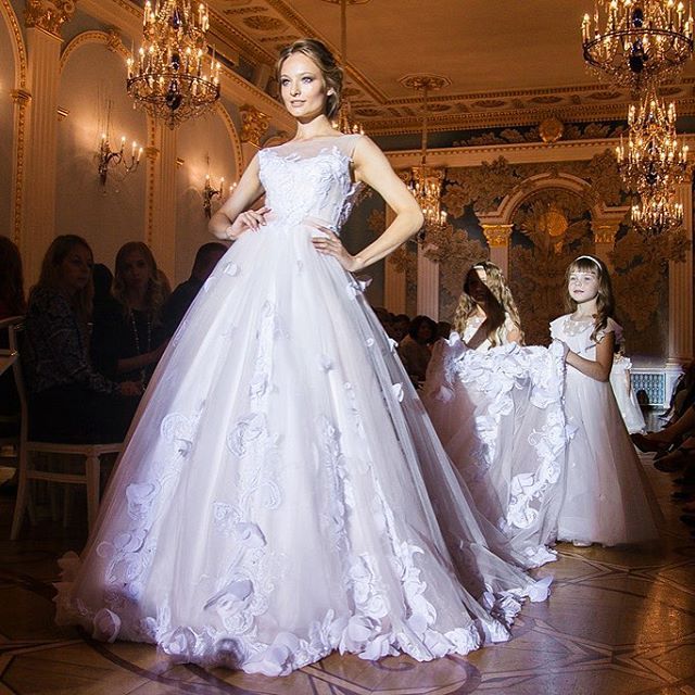 Woman in a white ball gown with floral appliqués and a fitted bodice, standing on a runway with two girls in white dresses behind her