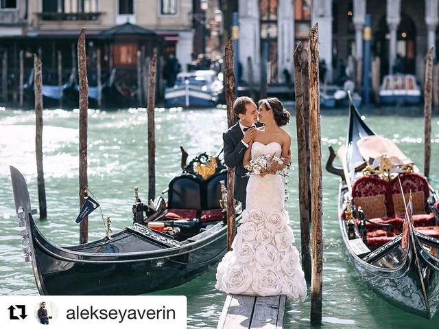 Woman in a white strapless wedding gown with floral appliqués, holding a bouquet, standing by gondolas on a canal