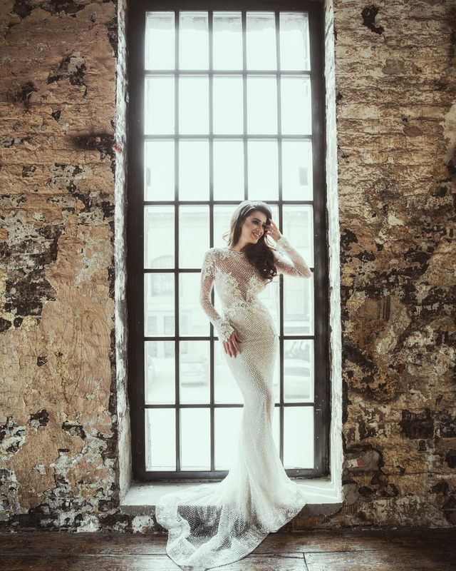 Woman in a white lace long-sleeve gown with a fitted silhouette and a flowing train, standing by a large window