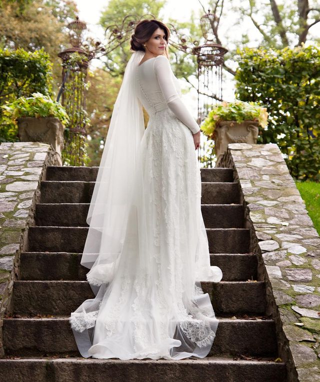 Woman in a long-sleeve white lace wedding gown with a flowing veil, standing on stone steps in a garden