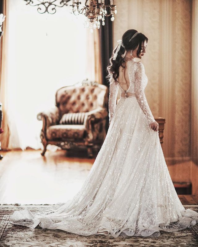 Woman in a white lace wedding gown with long sleeves and a flowing train, styled with loose waves and a ribbon in her hair