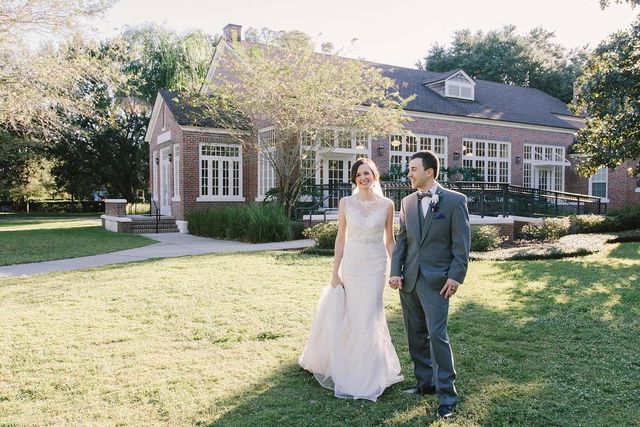 Woman in a white lace wedding dress and a man in a grey suit with a bow tie, holding hands outdoors