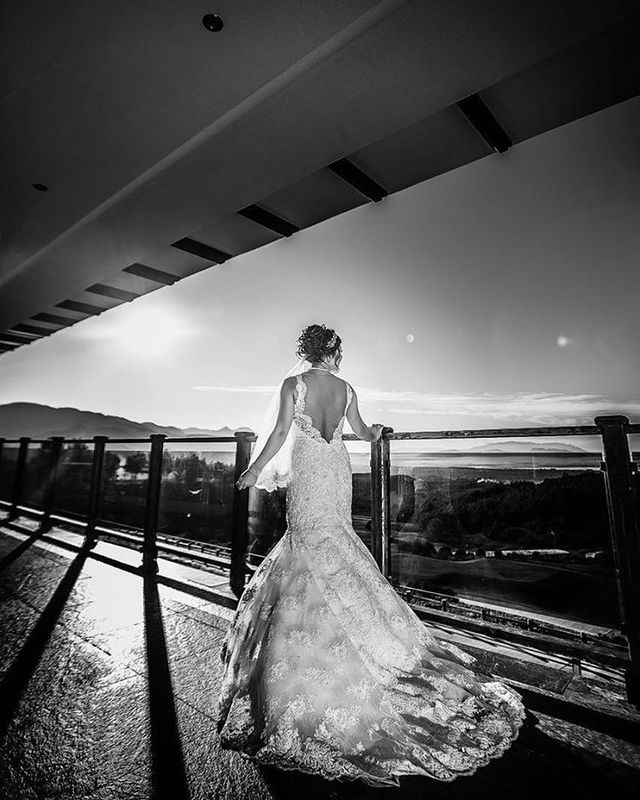 Woman in a white lace wedding gown with a fitted bodice and a flowing train, standing on a balcony at sunset
