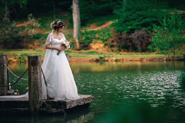 Woman in a white lace wedding gown with a flowing skirt, holding a bouquet of flowers by a lake