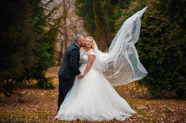 Woman in a white lace wedding gown with a full skirt and a long flowing veil, standing with a man in a black suit