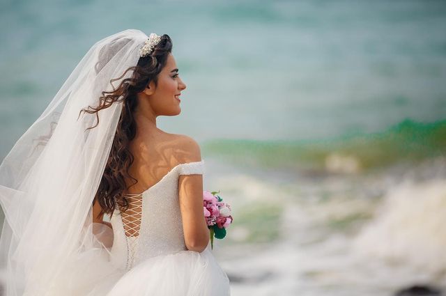 Woman in a white off-the-shoulder wedding gown with a lace-up back, holding a bouquet of pink flowers and wearing a long veil