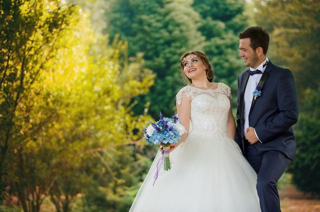Woman in a white lace wedding gown with a tulle skirt, holding a bouquet of blue flowers, and a man in a black tuxedo