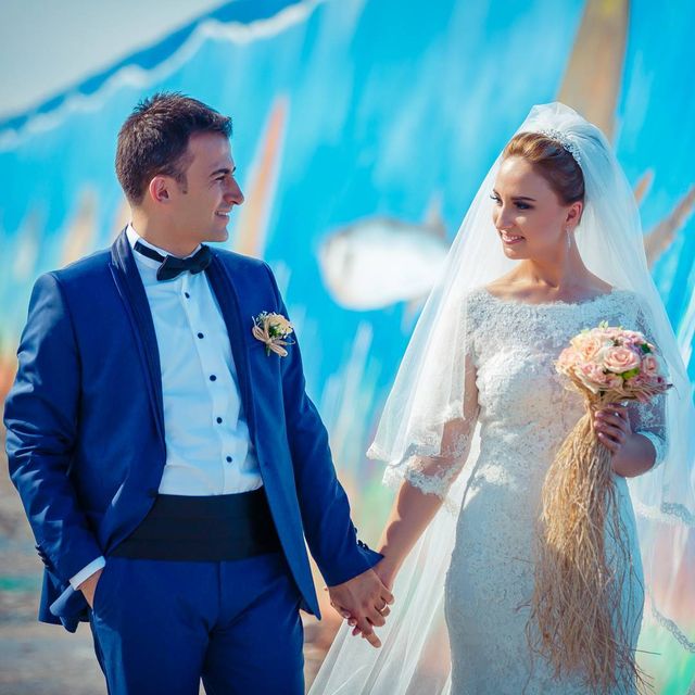 Woman in a white lace wedding dress with a long veil, holding a bouquet of pink flowers, alongside a man in a blue tuxedo
