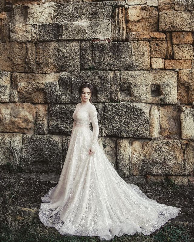 Woman in a long-sleeve ivory lace wedding gown with a flowing train against a stone wall