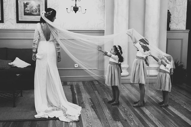 Woman in a white lace wedding gown with a long veil, accompanied by three girls in grey dresses and flower crowns
