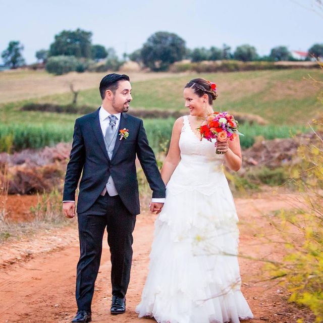 Woman in a white lace wedding dress with a tiered skirt, holding a colorful bouquet, walking with a man in a black suit and gray vest