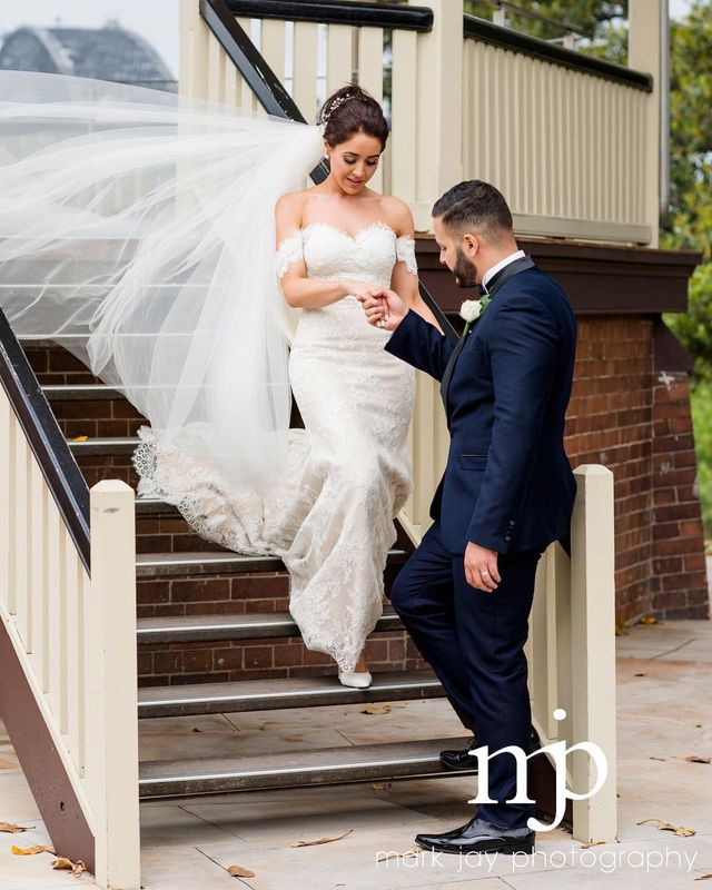 Woman in a white lace wedding gown with off-the-shoulder sleeves and a long veil, holding hands with a man in a navy suit