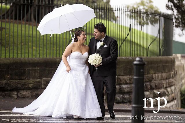 Woman in a white strapless wedding gown holding a bouquet and a white umbrella, walking with a man in a black tuxedo