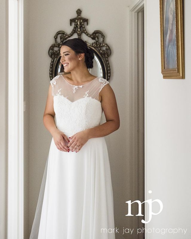 Woman in a white wedding dress with a sheer bodice and lace detailing, standing in a softly lit hallway