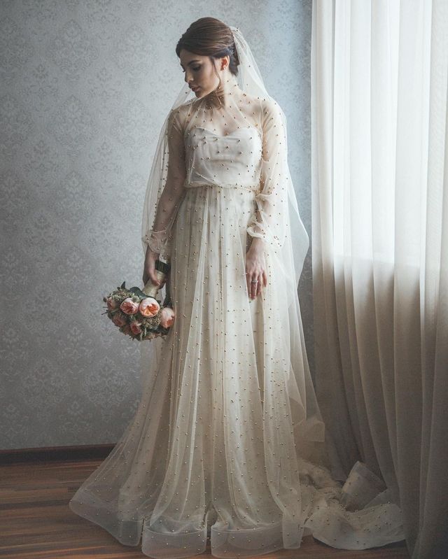 Woman in a sheer long-sleeve wedding gown with pearl embellishments and a flowing skirt, holding a bouquet of pink roses