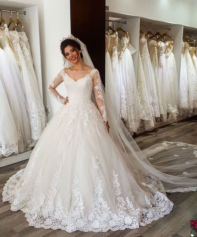 Woman in a white lace wedding gown with long sleeves and a flowing veil, standing in a bridal shop