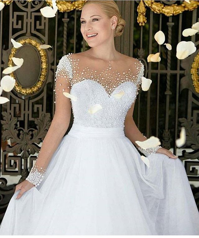 Woman in a white ball gown with a beaded bodice and sheer sleeves, holding the skirt with a floral backdrop
