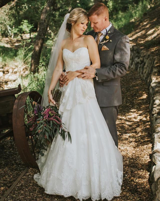 Woman in a white lace wedding gown with a sweetheart neckline, holding a bouquet of purple flowers and greenery