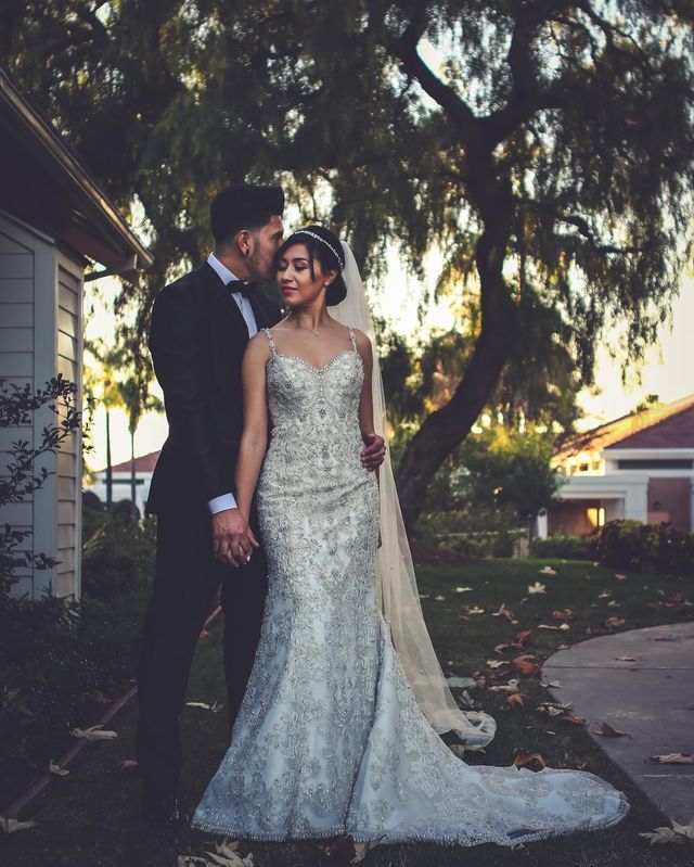 Woman in a white beaded wedding gown with a fitted silhouette and a long veil, standing beside a man in a black tuxedo