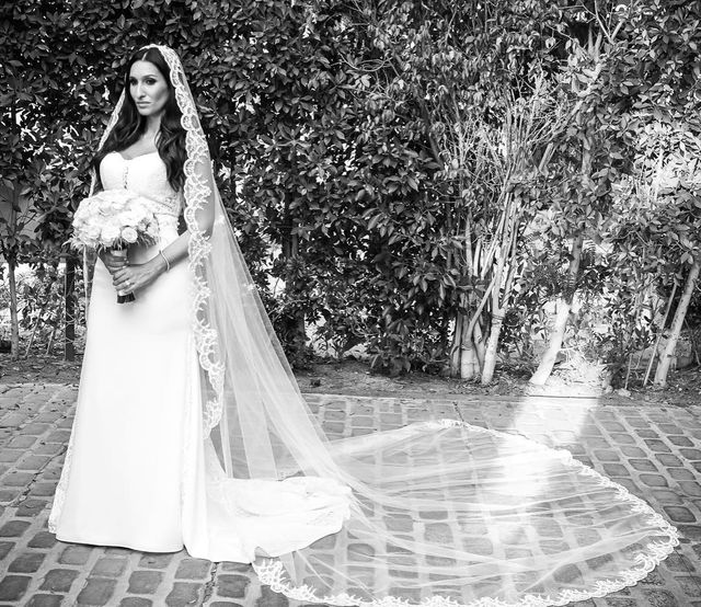 Woman in a white wedding gown with a fitted silhouette and a long lace-trimmed veil, holding a bouquet of flowers