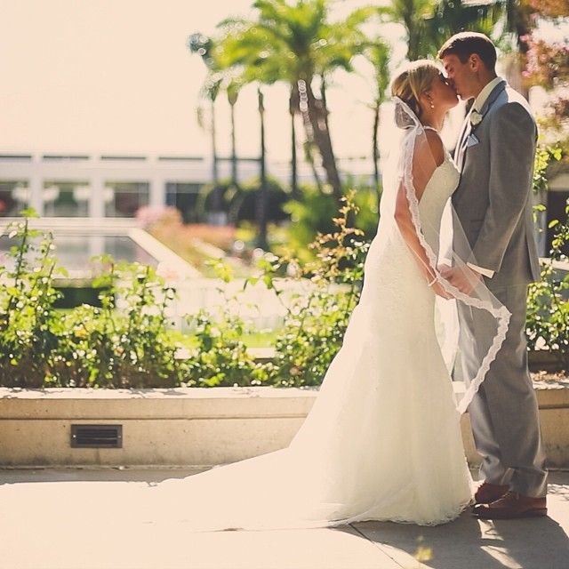 Woman in a white lace wedding gown with a long veil and a grey suit, standing outdoors among greenery
