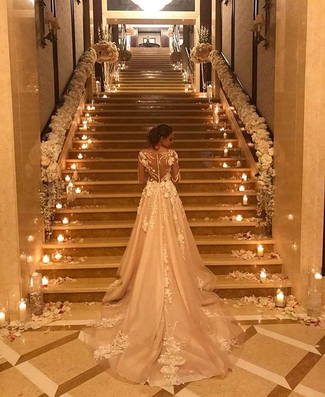 Woman in a nude lace wedding gown with a long train and sheer long sleeves, standing on a staircase adorned with candles and flowers