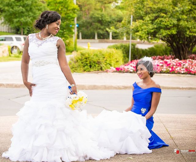 Woman in a white mermaid wedding gown with lace detailing and a pearl necklace, holding a bouquet of yellow flowers, alongside a woman in a blue off-the-shoulder dress with a fascinator