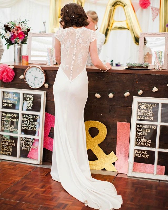 Woman in a white lace-back wedding gown with a fitted silhouette and a train, standing at a bar
