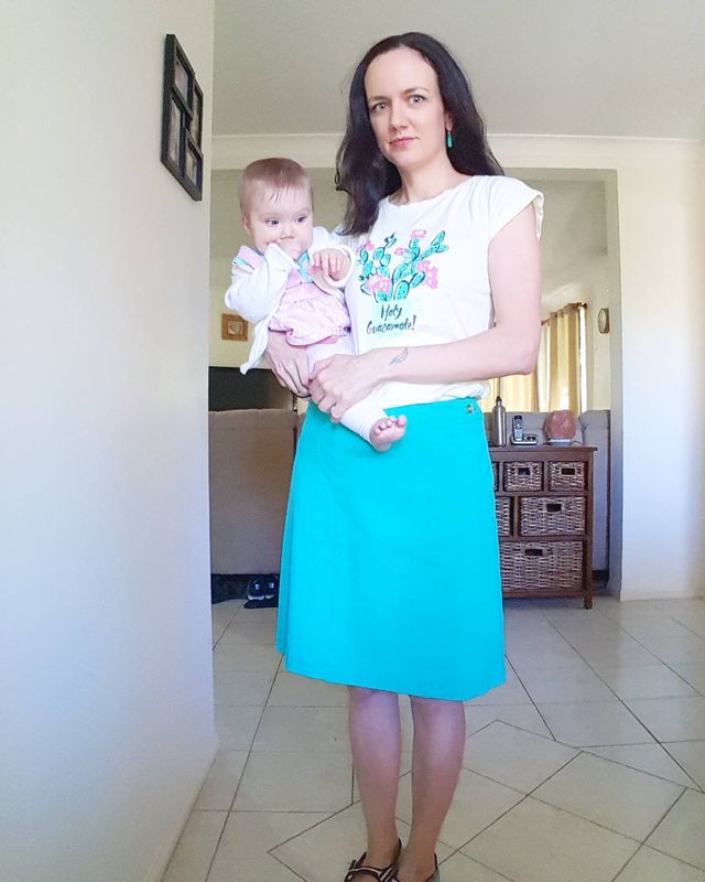 Woman wearing a white graphic t-shirt, turquoise knee-length skirt, and black sandals while holding a baby