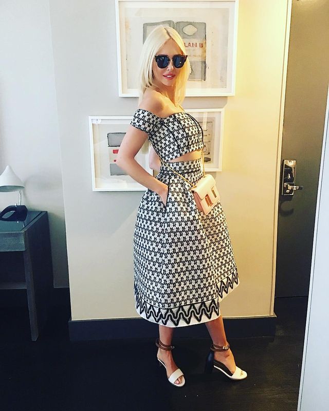 Woman in a black and white patterned off-the-shoulder crop top and matching midi skirt, with white strappy heels and sunglasses