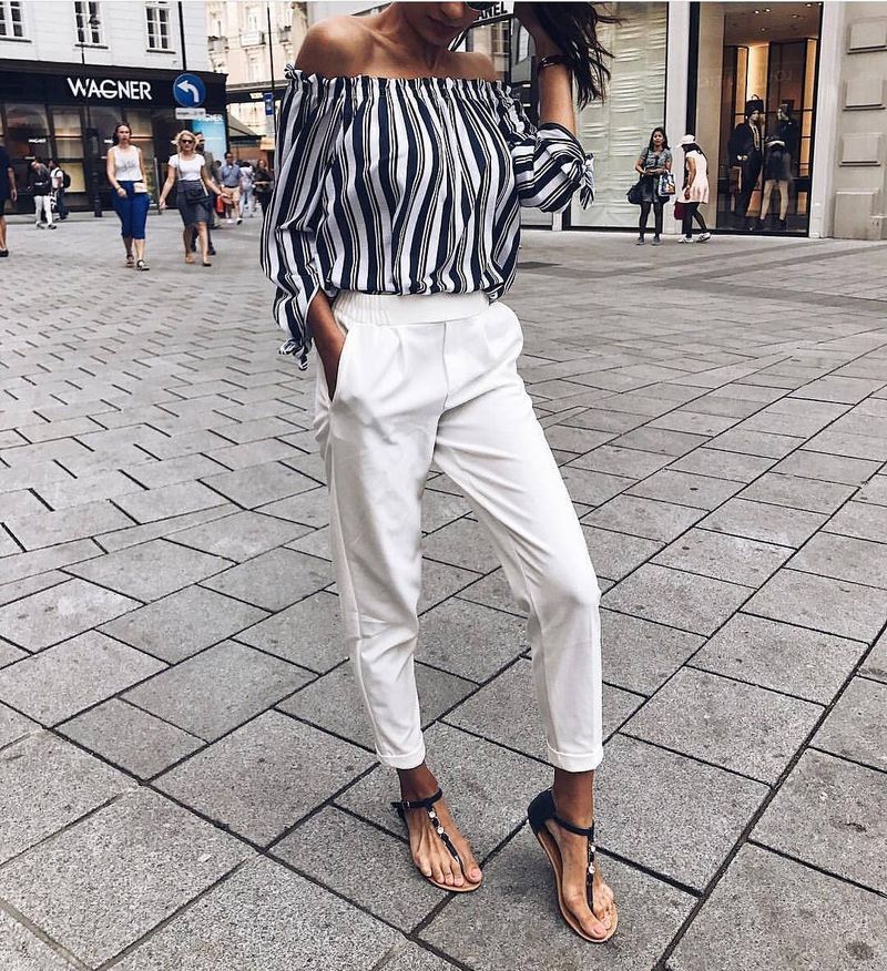 Woman wearing a striped off-the-shoulder blouse, white tailored trousers, and black flat sandals on a city street