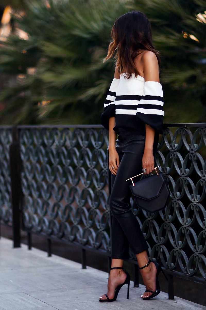 Woman wearing a black and white striped off-the-shoulder top, black leather pants, and black strappy heels, holding a black handbag