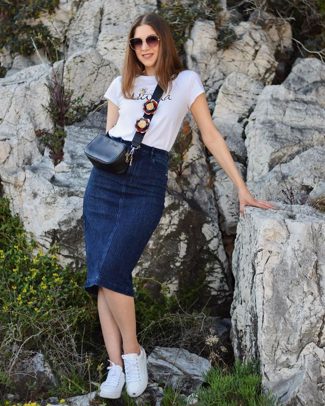 Woman wearing a white graphic t-shirt, dark denim pencil skirt, and white sneakers with a black crossbody bag and sunglasses