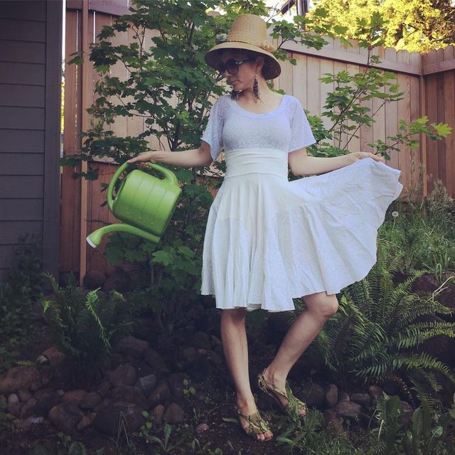 Woman wearing a white sundress, sheer white top, and tan wedge sandals, holding a green watering can in a garden setting